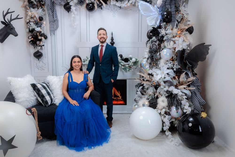 Elegant couple in blue dress and suit embrace near decorative white and black holiday decor.