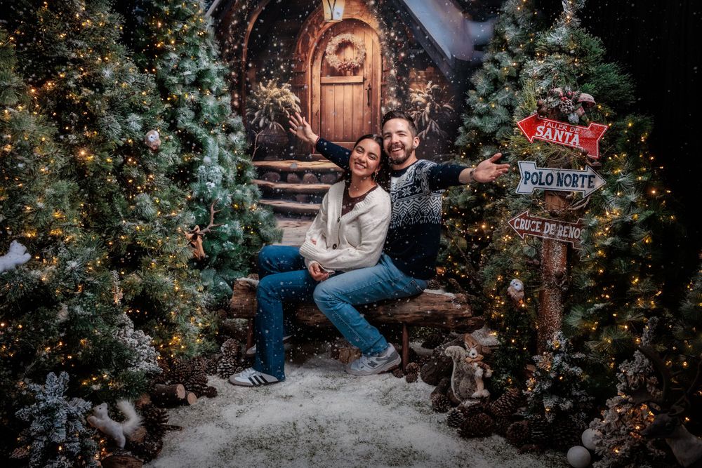 Couple poses in a cozy winter wonderland setting with twinkling lights and evergreen trees.