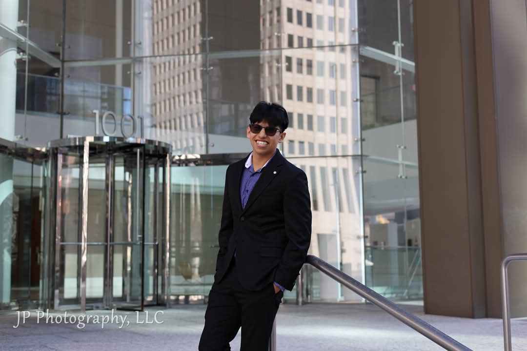 Professional portrait of a person in a dark suit standing in front of a modern glass building.