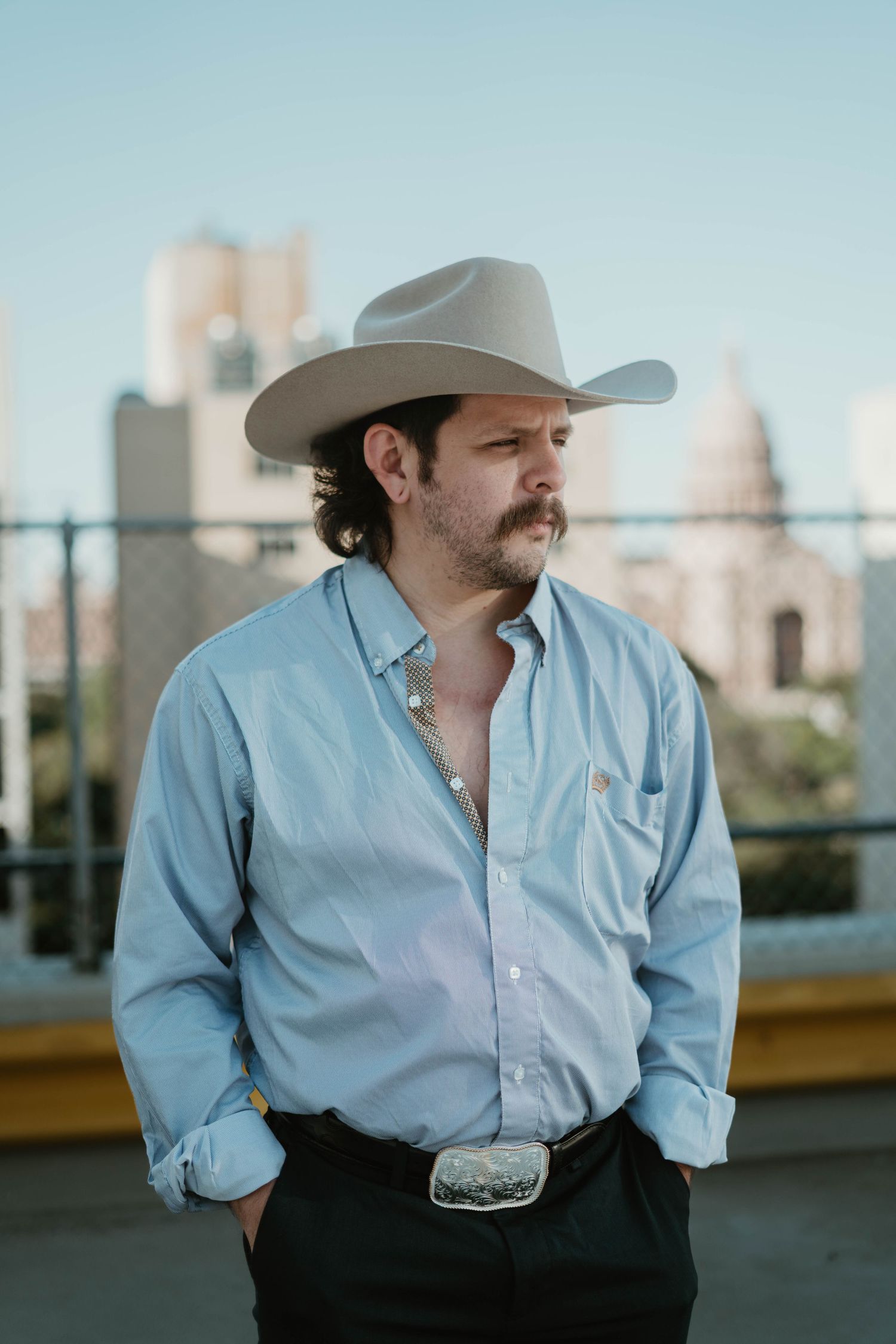 Person in blue button-down shirt and cowboy hat poses against urban backdrop with rolled sleeves and western belt buckle.