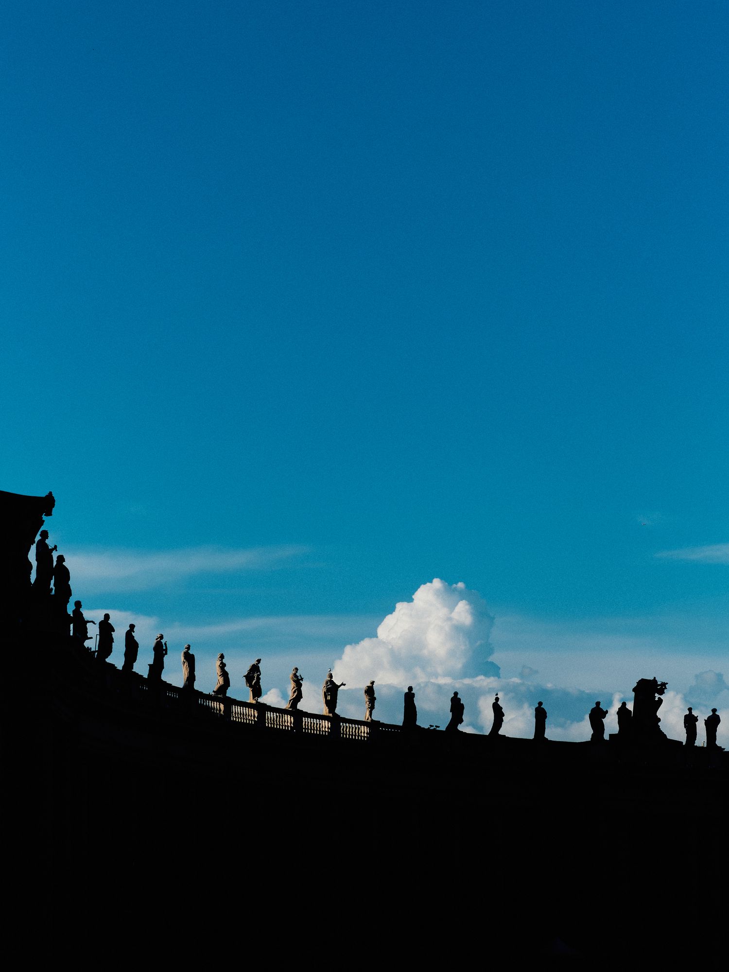 Silhouetted figures walking along ridge against bright blue sky with white puffy cloud in background.