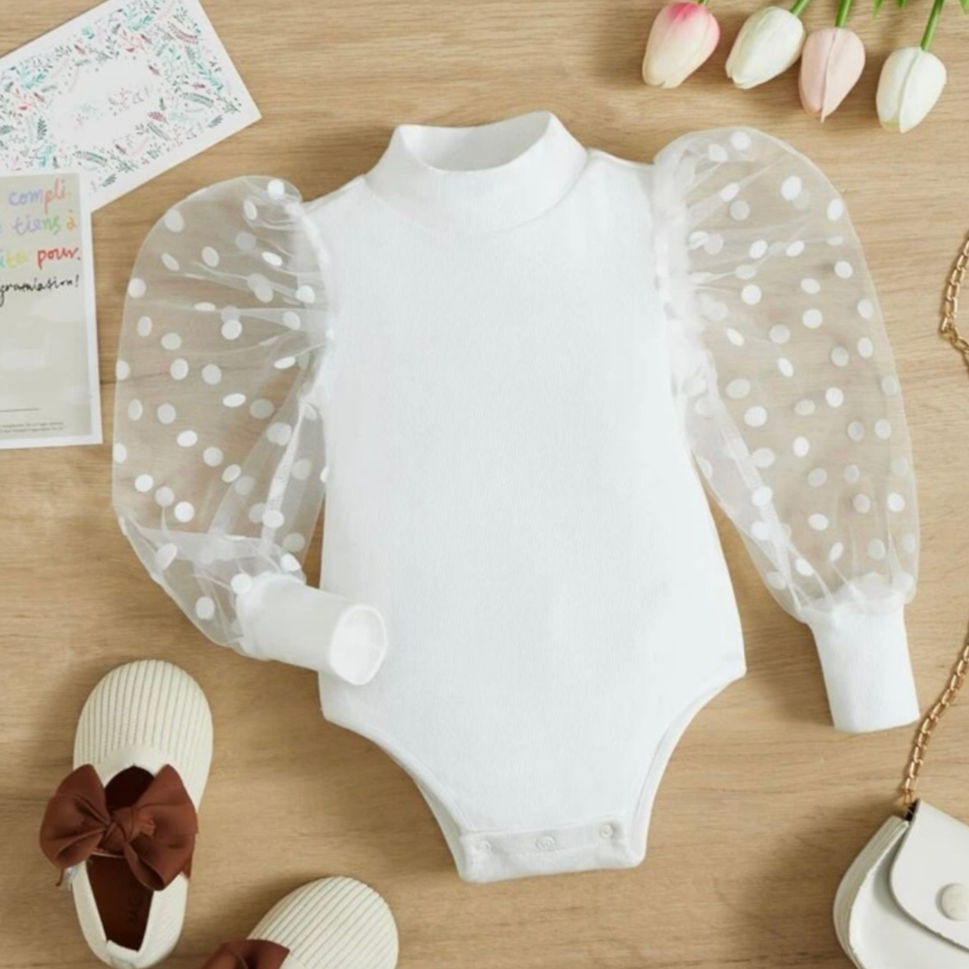 White polka dot baby onesies and clothing items arranged on a wooden surface with pink tulips.