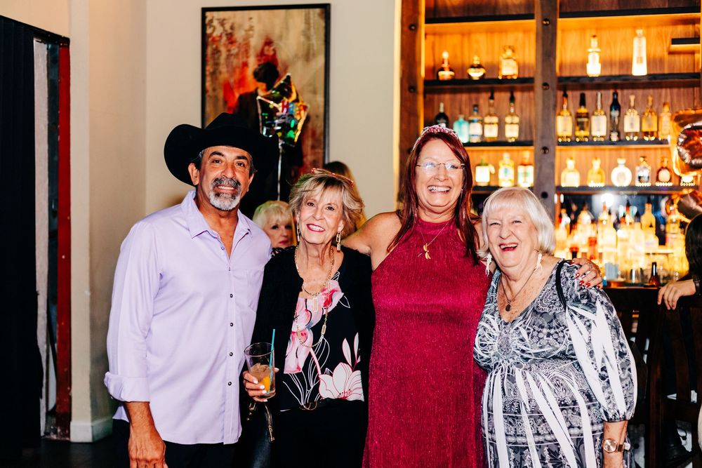 Friends gather for a group photo at a bar with liquor bottles and warm lighting in the background at an event in lake worth.