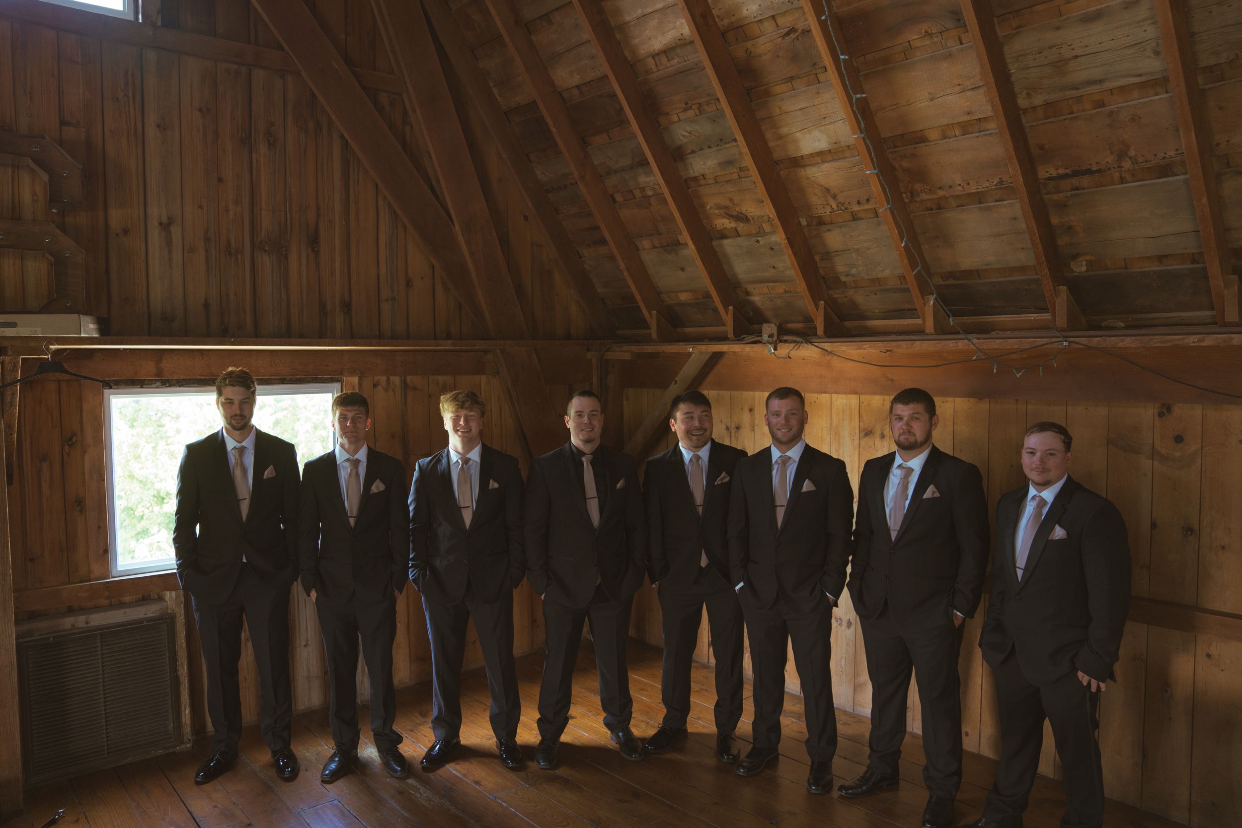 Wedding party groomsmen lined up in formal attire inside rustic wooden barn.