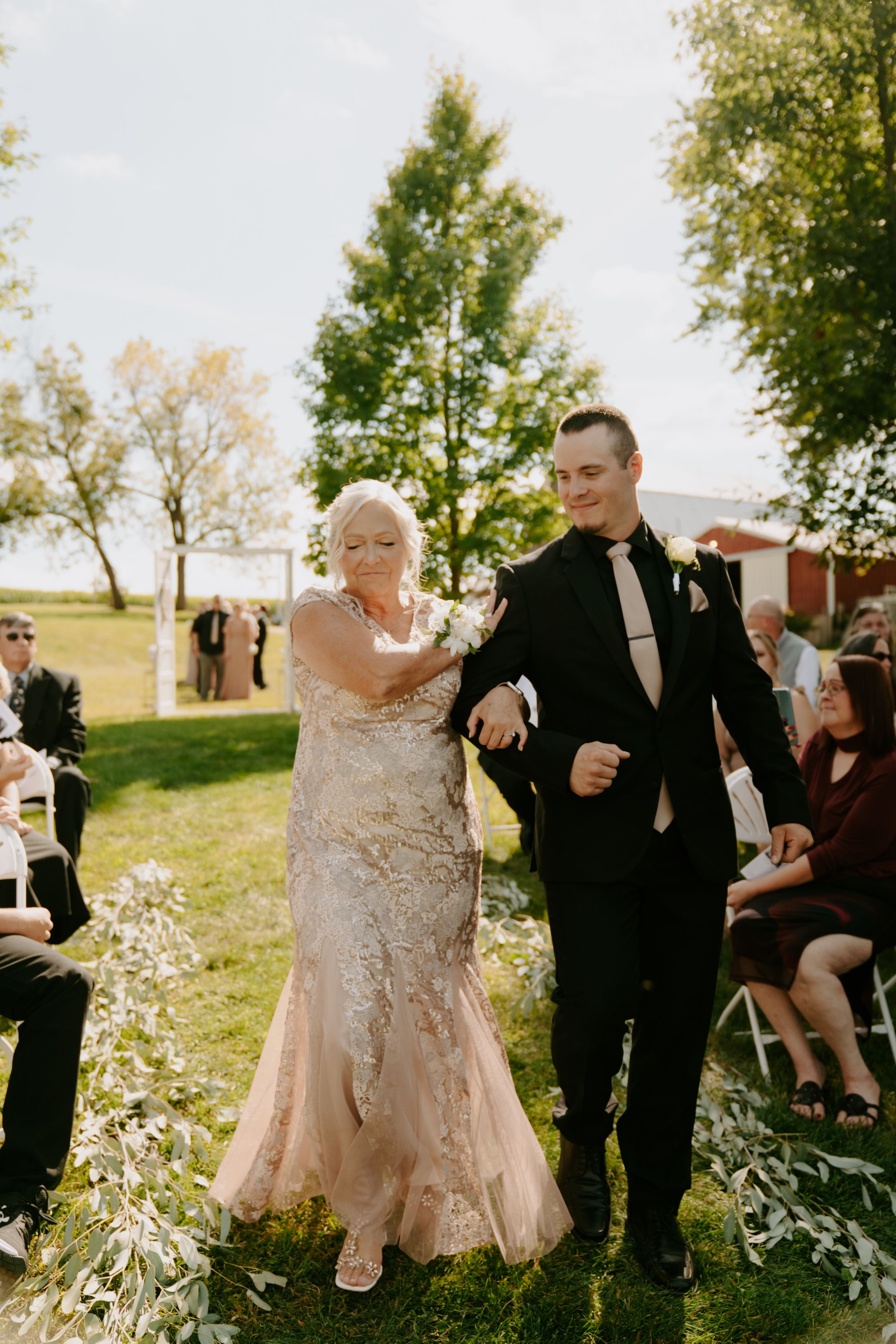 Outdoor wedding processional with attendants walking through garden setting.