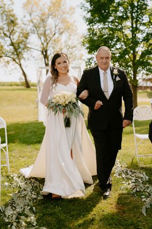 Elegant outdoor wedding processional with flowing white dress and greenery decorations.