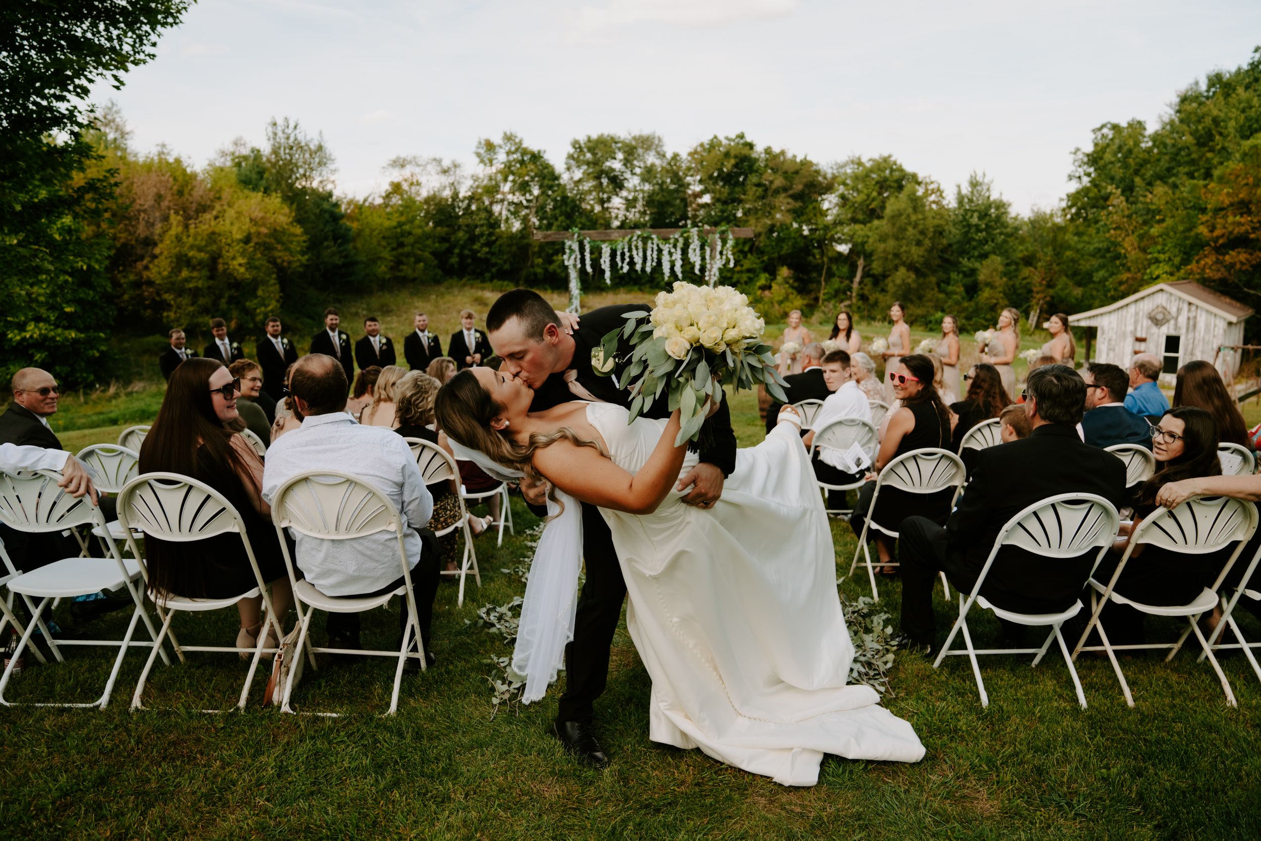 Wide angle view of outdoor wedding ceremony with guests seated in white folding chairs.