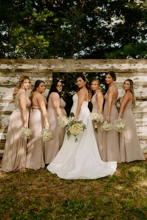 Wedding party in neutral dresses poses against stone wall in outdoor garden setting.