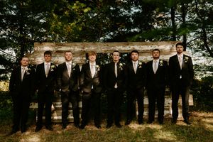 Groomsmen in black suits and ties stand in line against rustic wall backdrop.