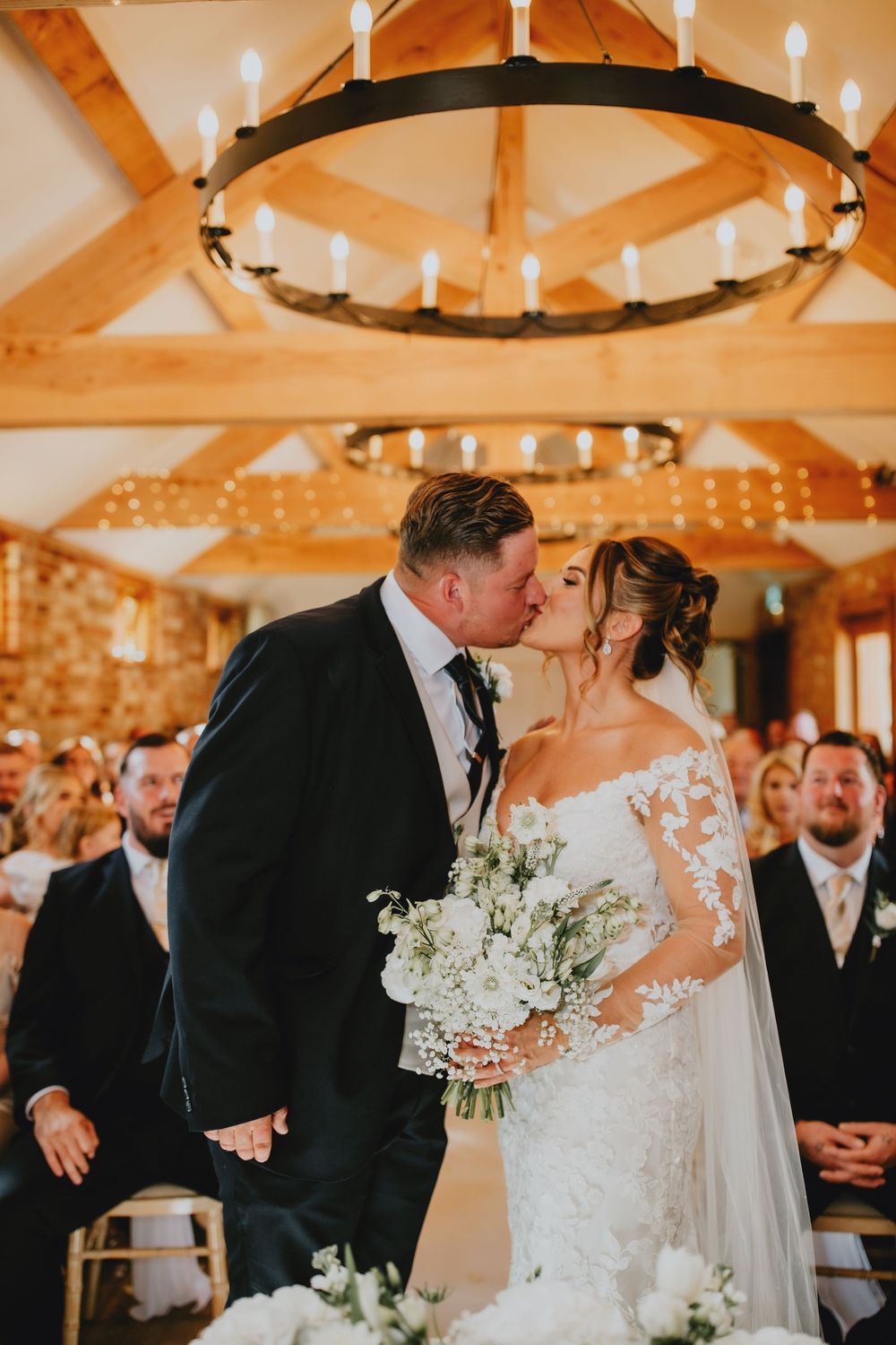 Couple sharing their first kiss during wedding ceremony under rustic wooden beams with chandelier and string lights.