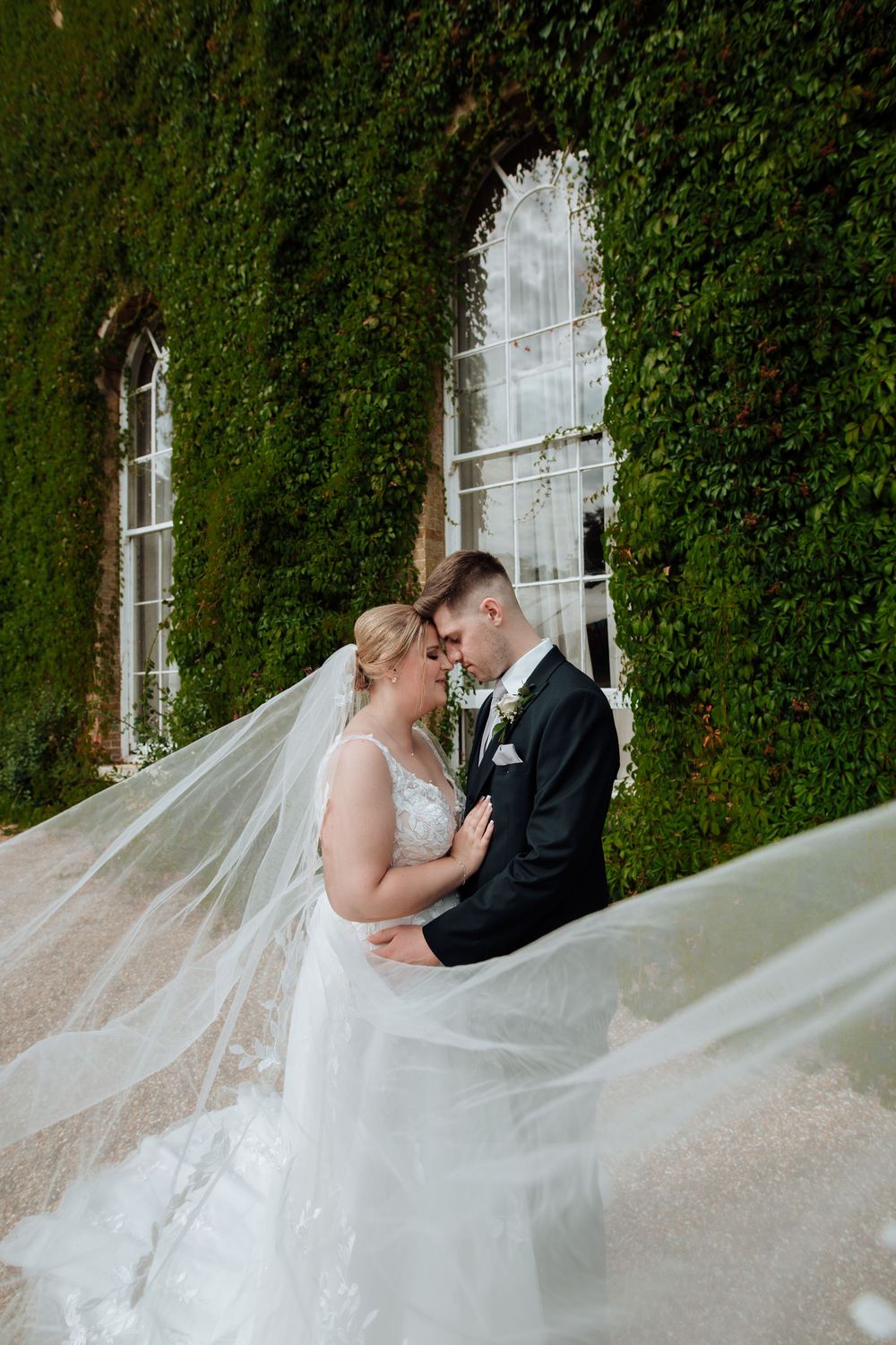 Newlyweds embrace in a romantic moment against an ivy-covered wall with arched windows in an elegant garden setting. De Vere Beaumont Estate