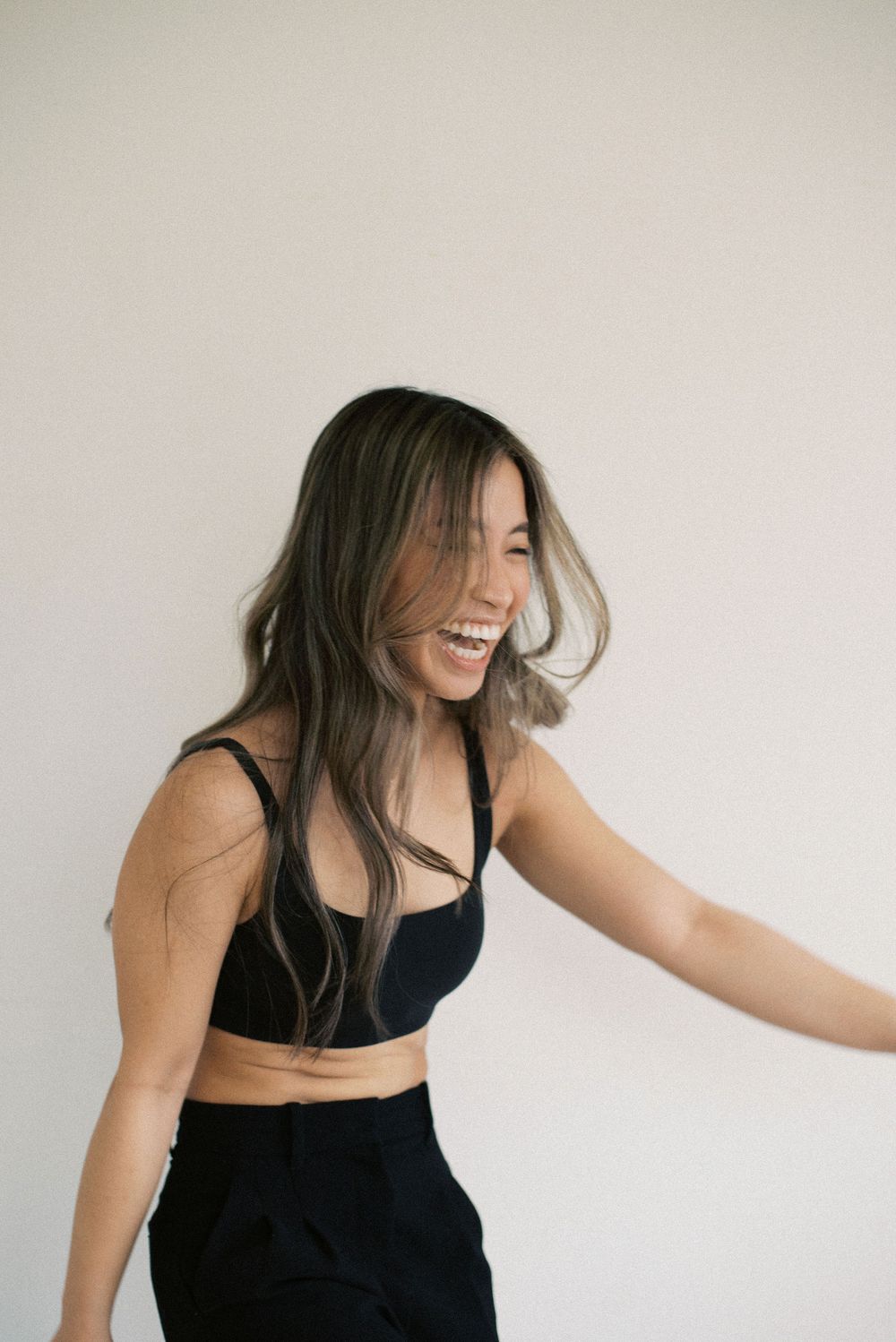 Woman in black crop top and pants laughing joyfully against white wall.