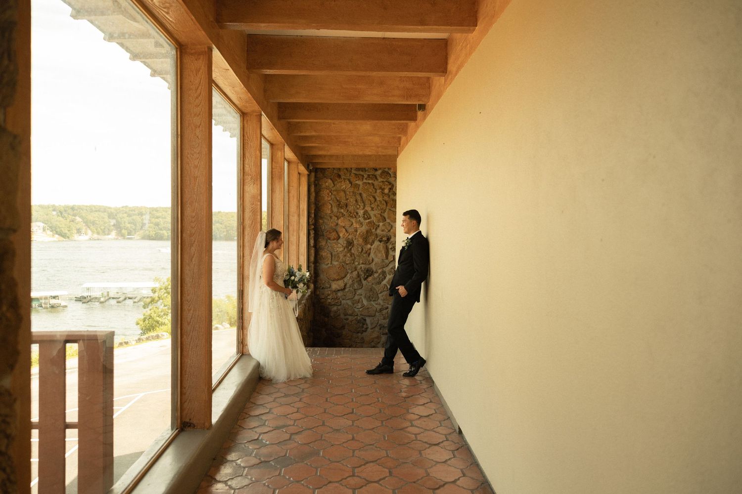 A bride and groom stand in a sun-lit hallway with stone walls and wooden beams overlooking a scenic waterfront view.