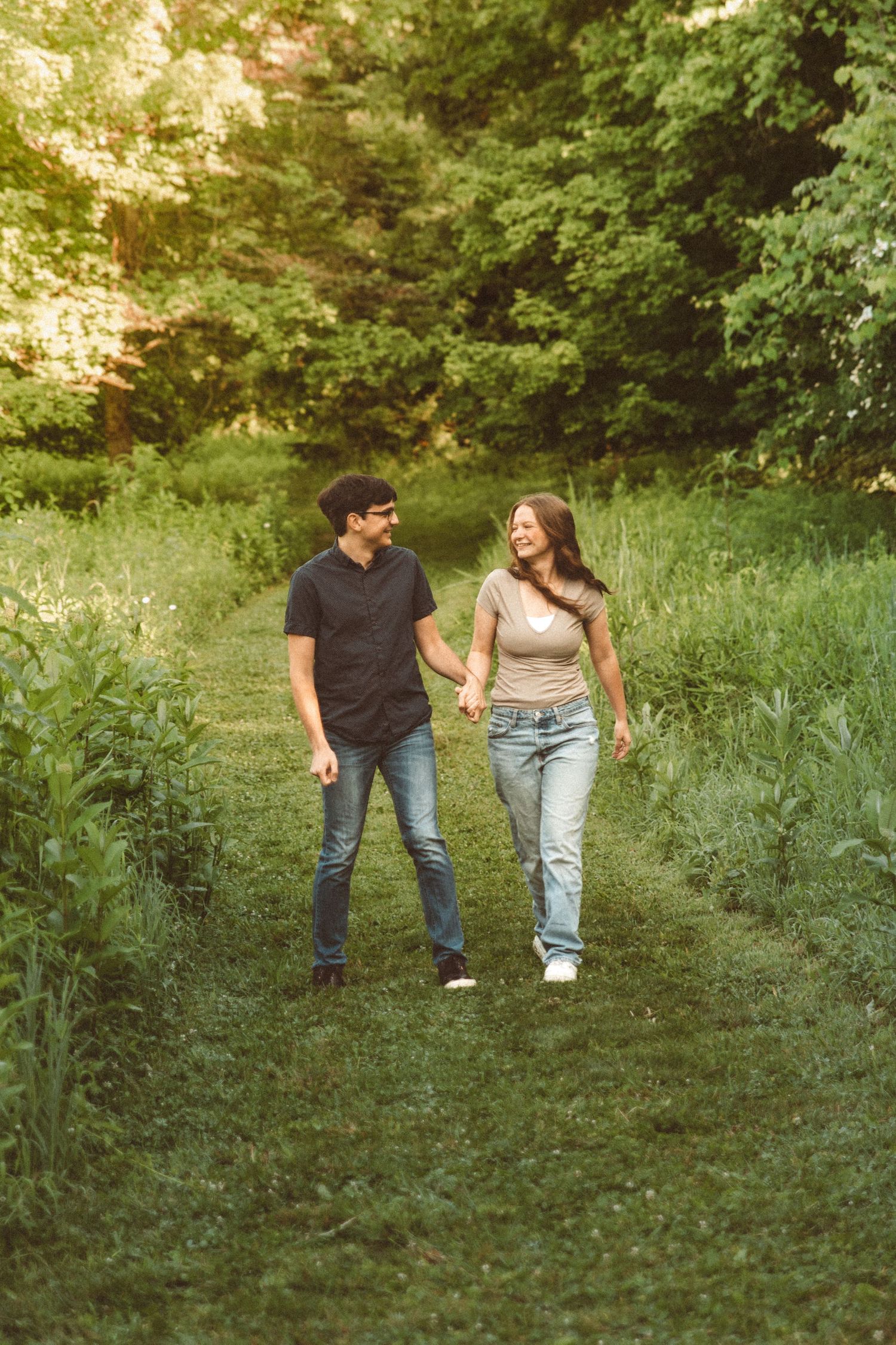 A young couple walking through a grassy path smiling at each other.