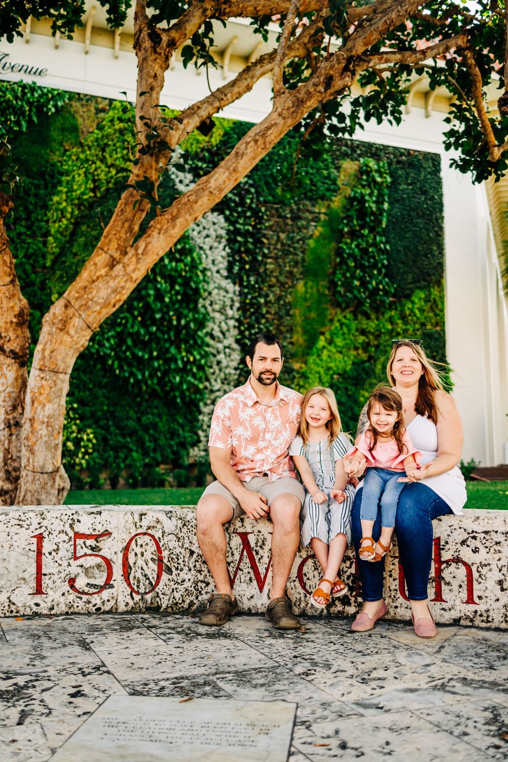 Family sitting together on stone wall with '150 Worth' text in tropical garden setting at worth ave palm beach Florida in palm beach Florida