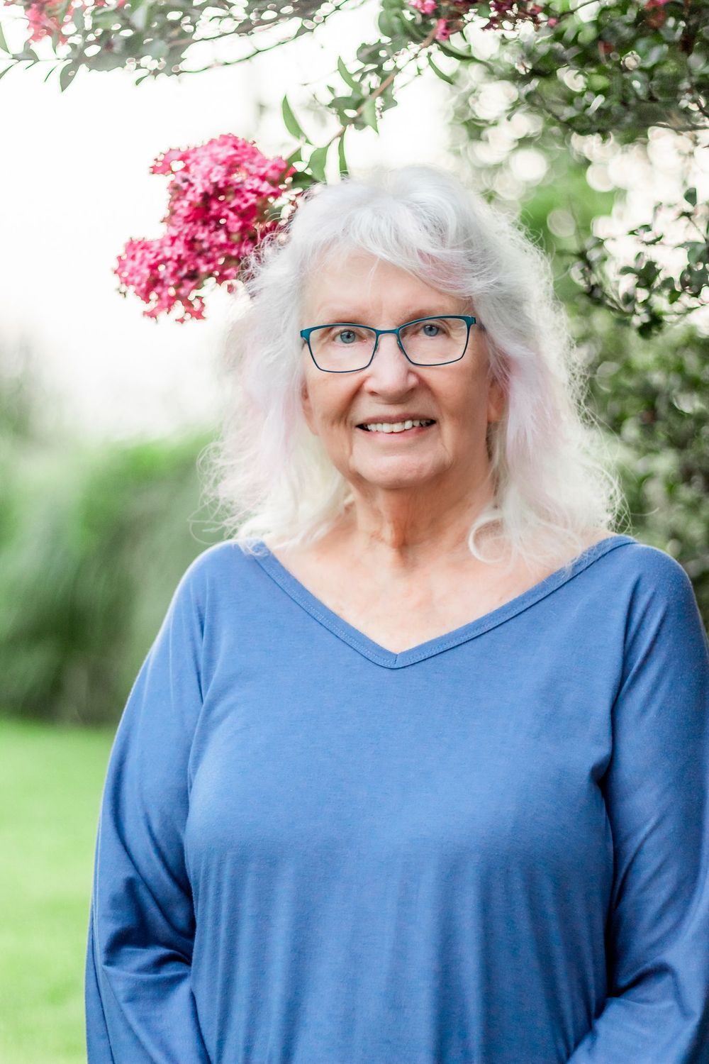 A grandmother in blue top smiling warmly in front of pink flowering bushes.