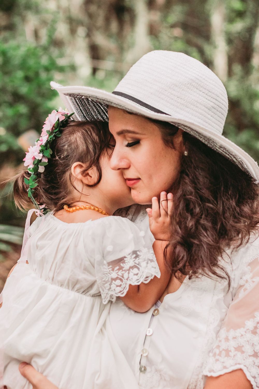 Mother in white sun hat embraces child wearing floral crown in ethereal outdoor portrait.