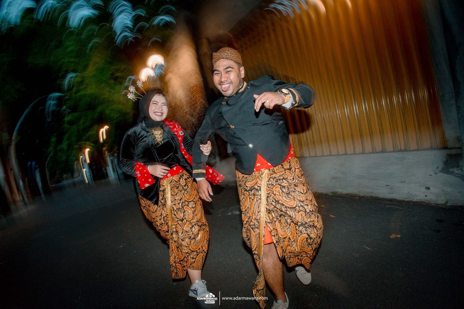 Two people wearing traditional Indonesian batik sarongs dance energetically in a dimly lit walkway or tunnel.