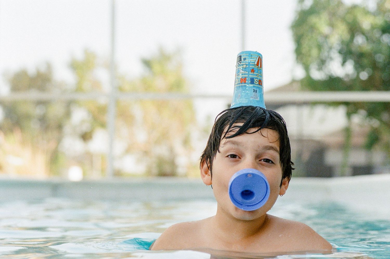Young child wearing blue nose plug plays in swimming pool with castle toy balanced on head.