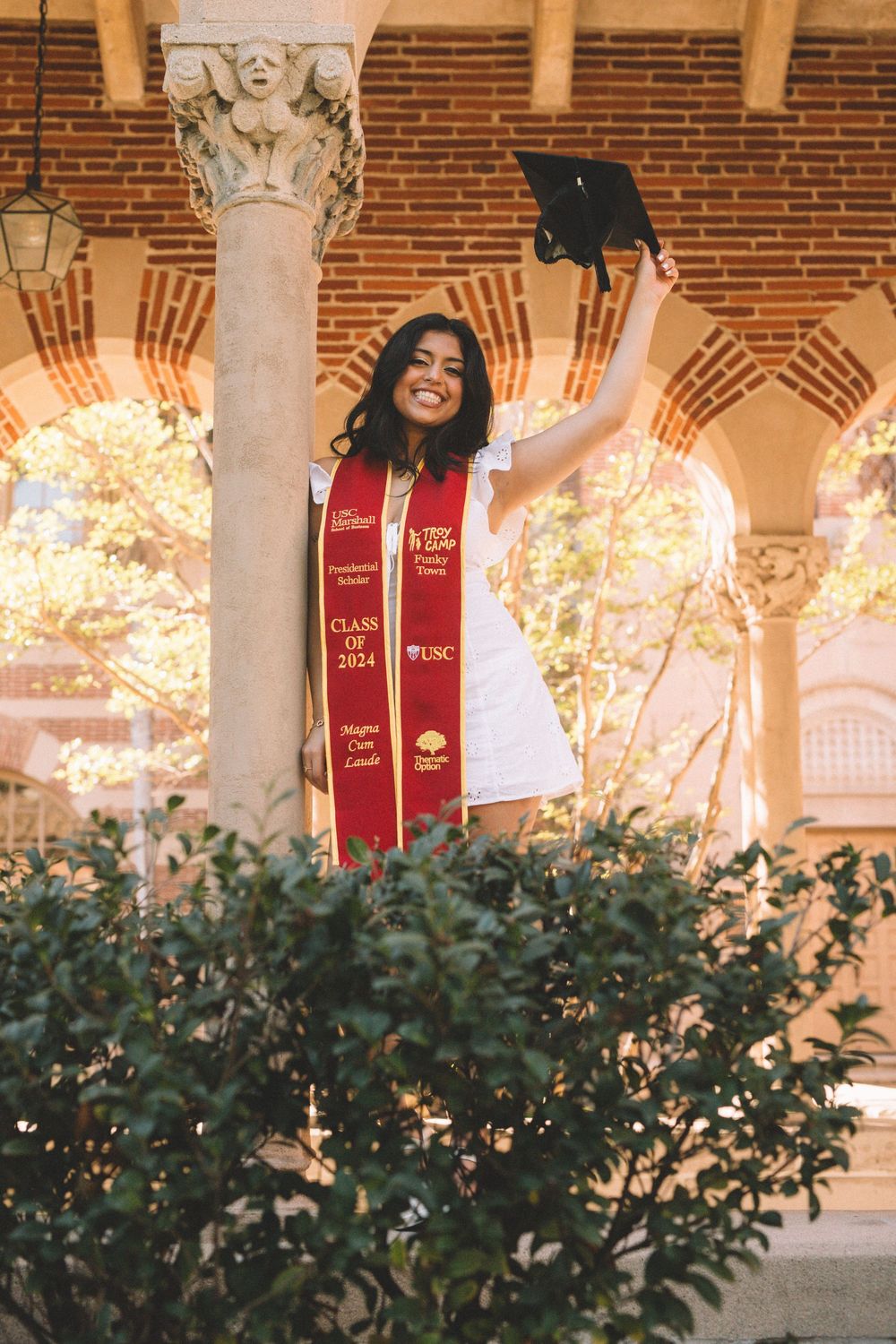 Graduate in white dress and red stole celebrates by holding cap up near ornate column.