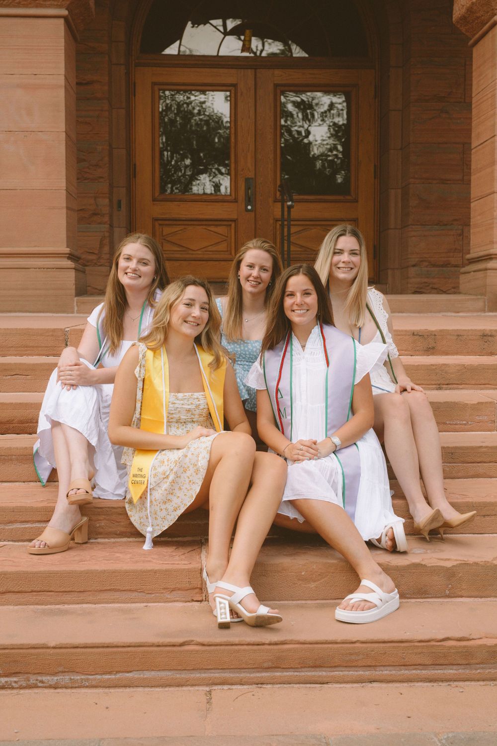 Five graduates in white and pastel dresses sit on stone steps.