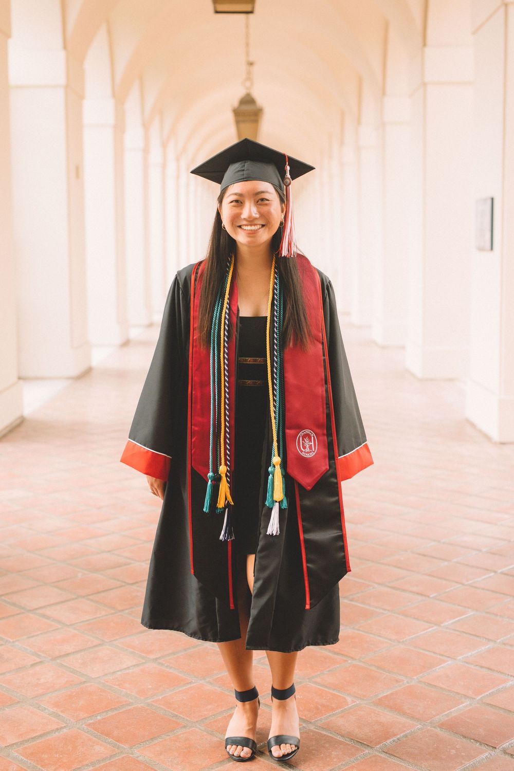 Graduate wearing black cap and gown with red stole stands in arched hallway.