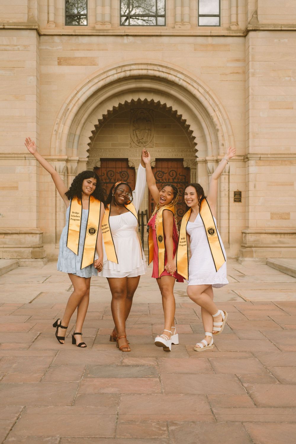 Four graduates in white dresses and gold stoles celebrate under arched doorway.
