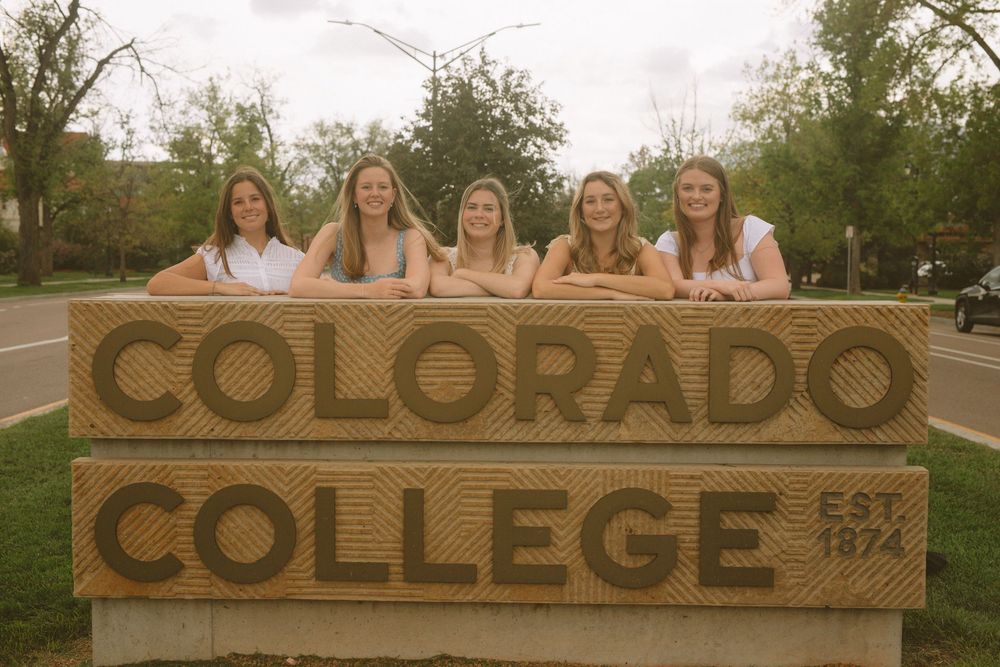 Five graduates in white dresses pose behind wooden Colorado College sign.