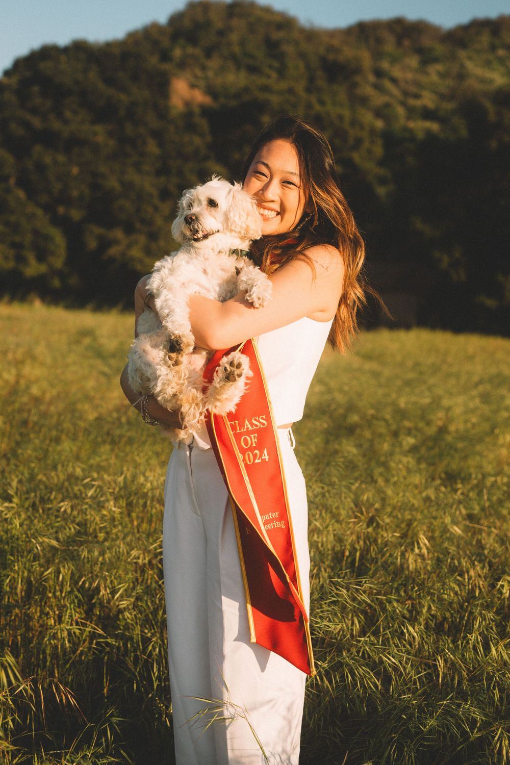 Graduate in white dress and red sash holds small white dog in sunny outdoor setting.
