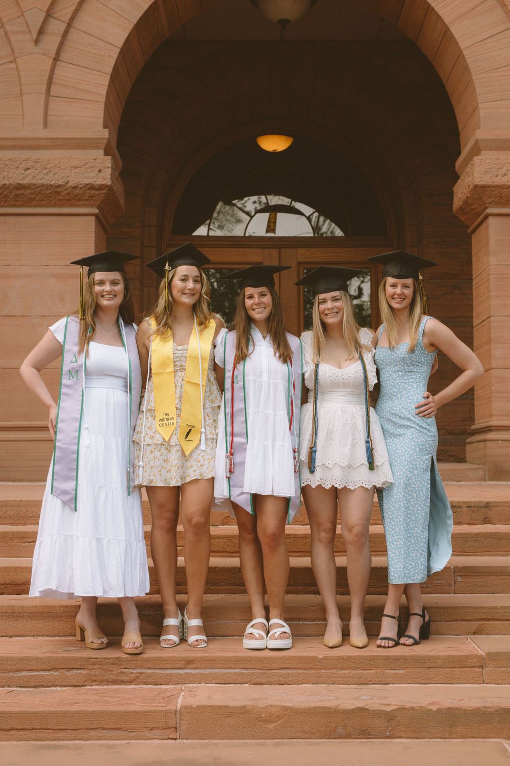 Five graduates in light dresses and stoles pose on steps of historic building.