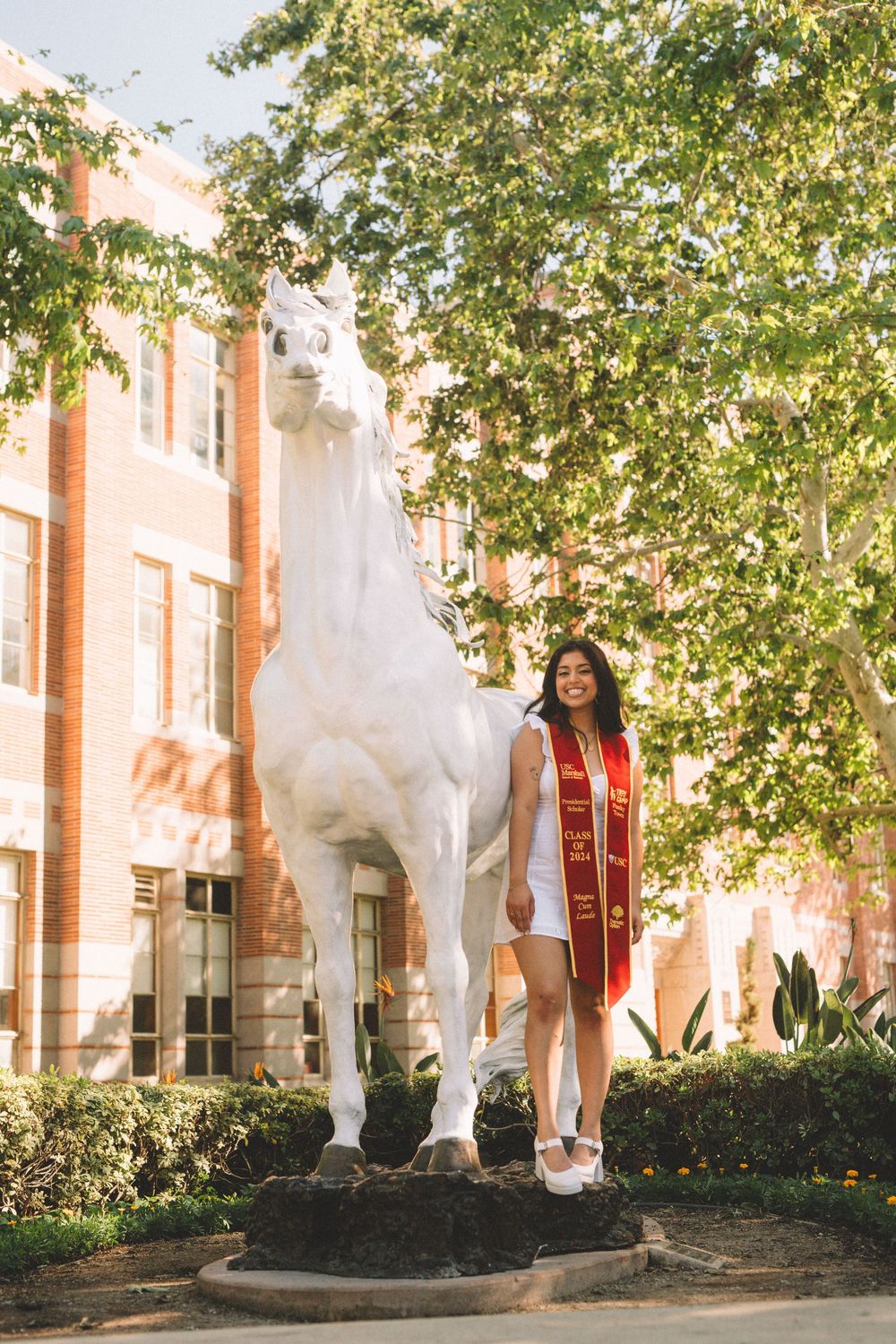 Graduate in white dress and red stole poses next to white horse statue on campus.