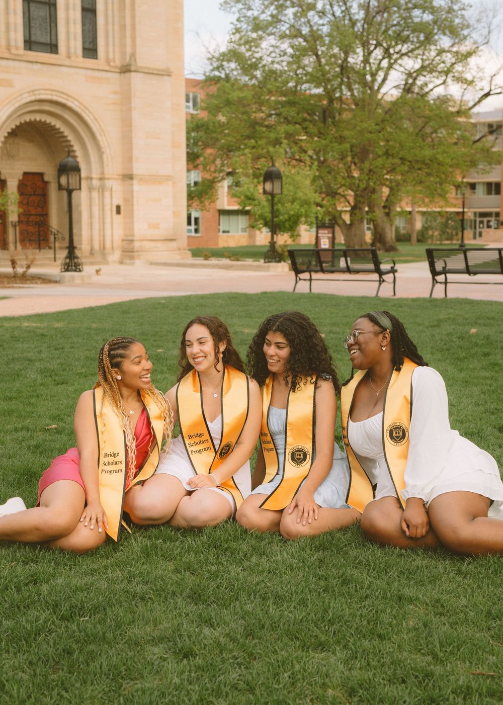 Four graduates in white dresses with gold stoles relax on campus lawn.