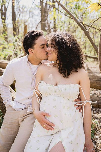 A couple in white and beige outfits share tender moments in a wooded outdoor setting during a romantic photoshoot.