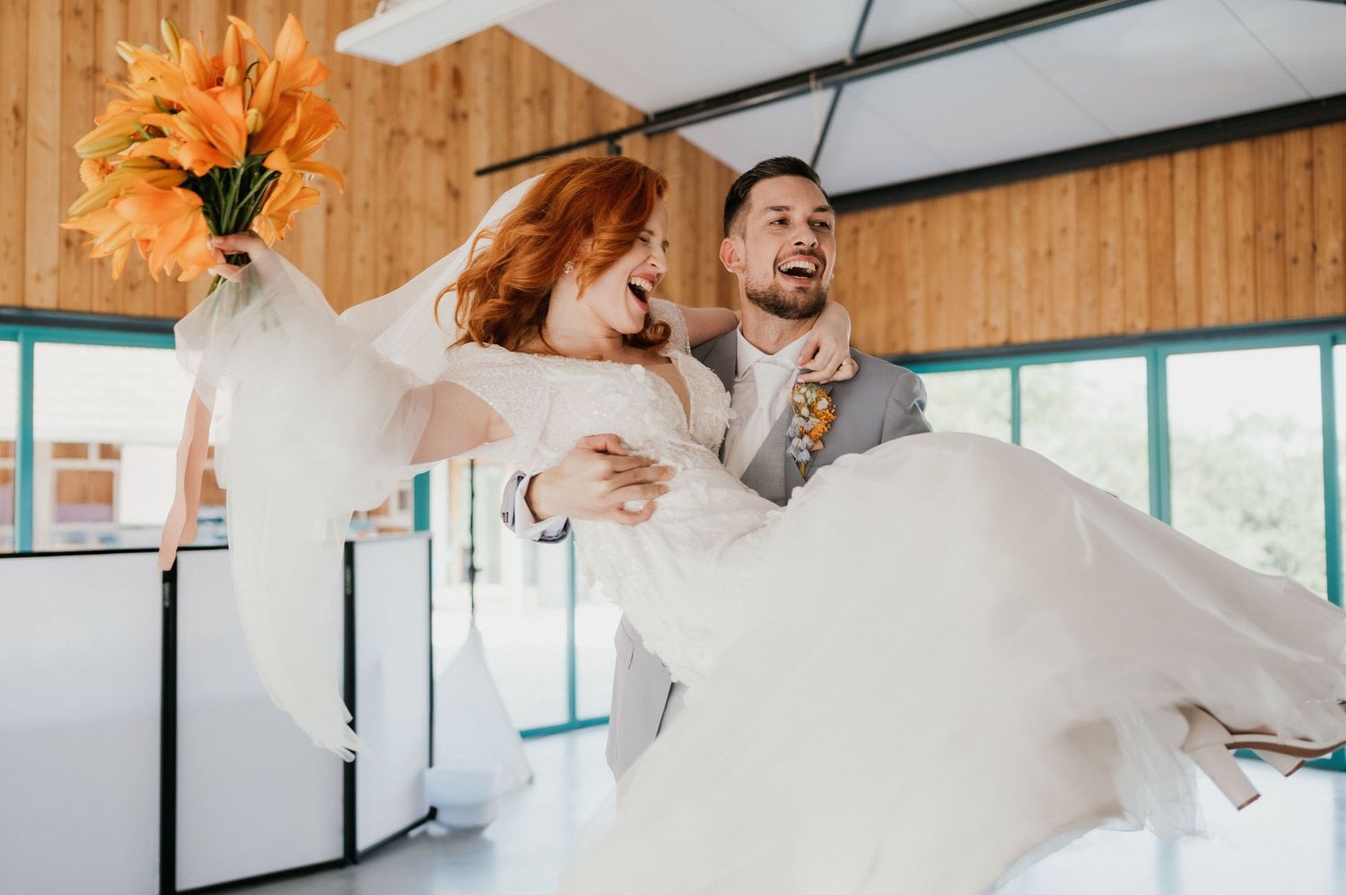 A joyful wedding moment showing a couple laughing while during a celebratory carry in a wood-paneled venue.