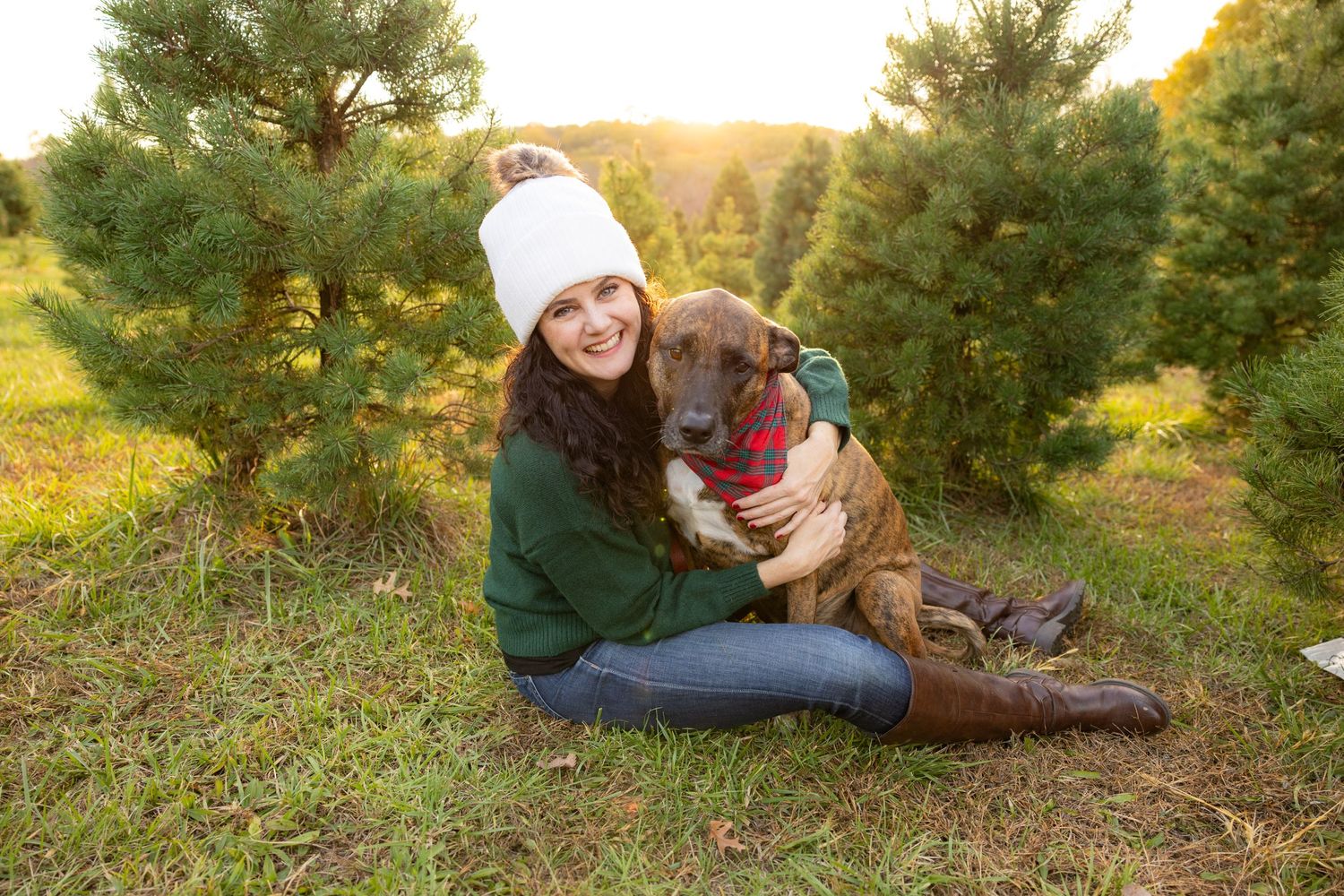 A cozy winter scene among evergreen trees shows companionship between a boxer dog and someone in a white beanie and green sweater.