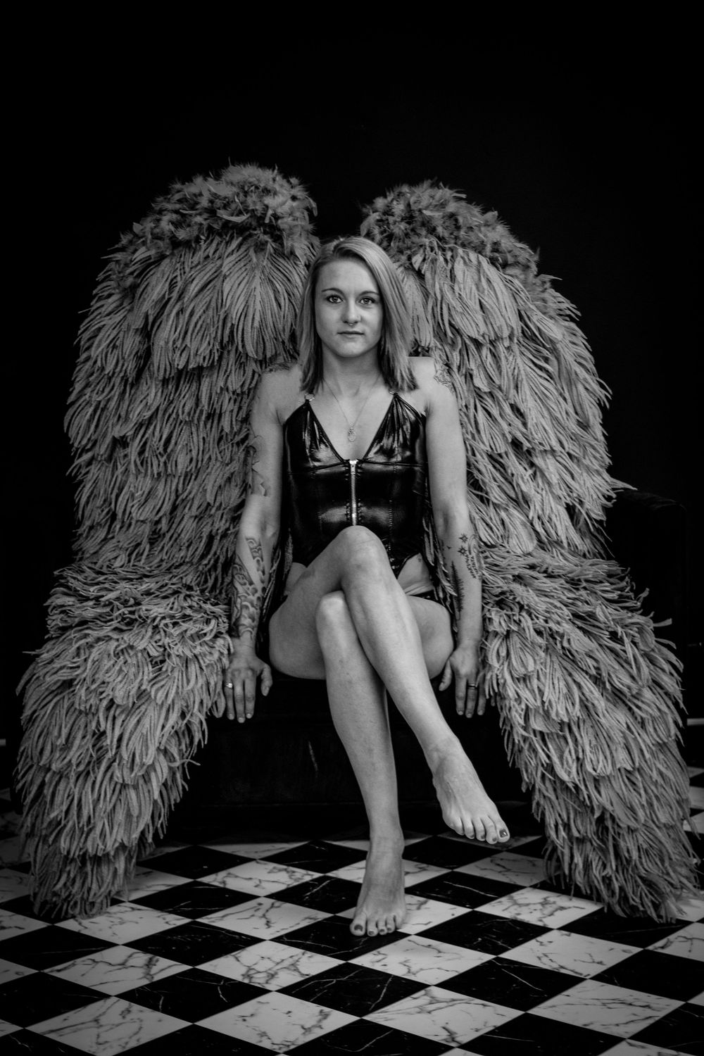 Black and white portrait of someone in a black dress posed with large feathered wings on a checkered floor.