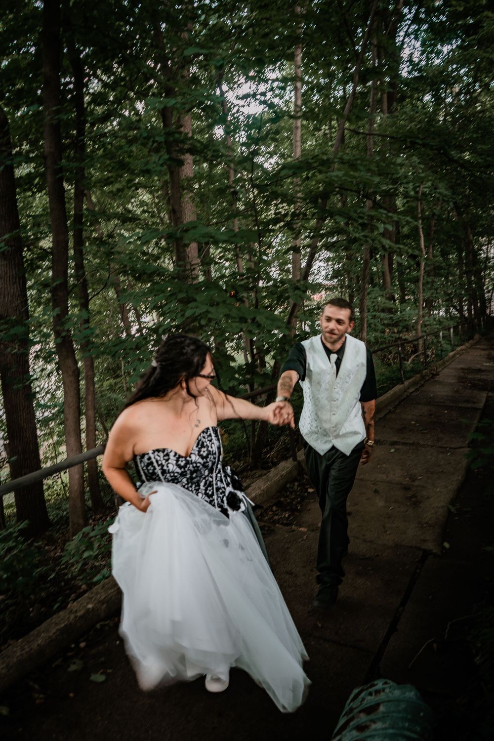 Couple in wedding attire walking along wooden boardwalk in forest setting.