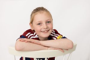 Young student rests their arms on a white chair while wearing a striped sweater against a light background.