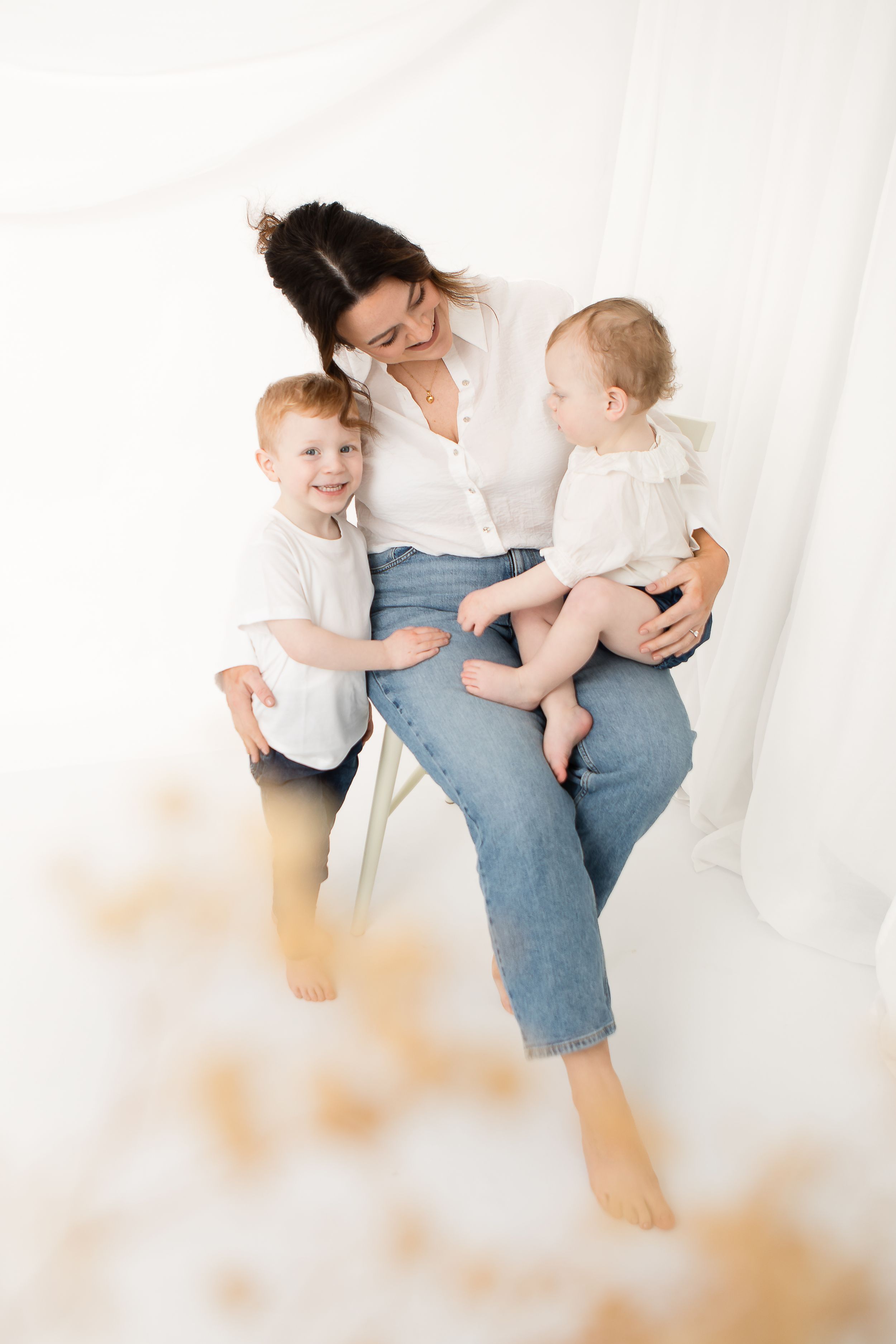 Family portrait of mother in casual denim and white top sitting with two young children against light background.