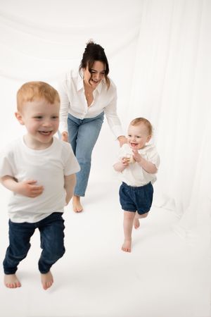 Joyful children in white shirts and denim run barefoot on white background while adult in casual attire follows behind.