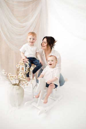 Three people in white clothing sit and smile together on a chair against a light draped backdrop with flowers.