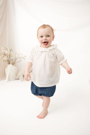 A happy baby in a white blouse and denim shorts stands against a white background with decorative flowers.