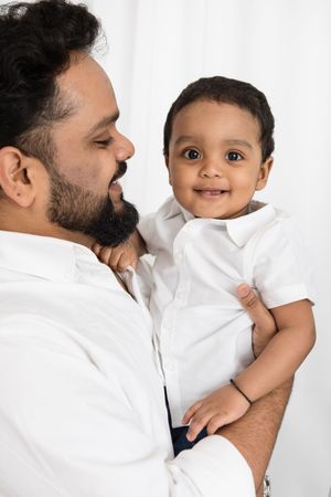 A person in a white shirt holds and smiles at a happy baby also wearing white against a light background.