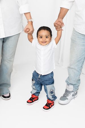 Small child wearing white t-shirt and jeans holds hands with adults while wearing red and black sneakers.