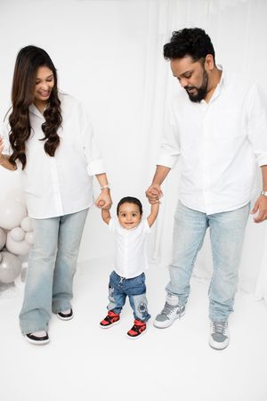 Family wearing coordinated white tops and light denim stands together on white background holding hands.