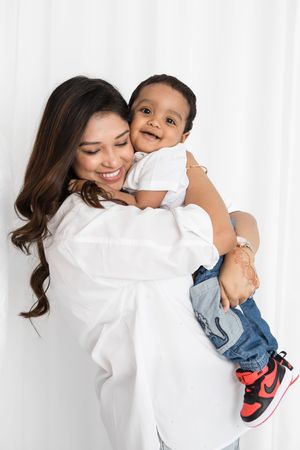 A woman in white embraces a young child wearing jeans and red sneakers against a white backdrop.
