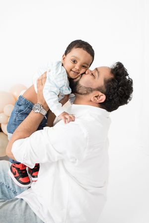 A father in a white shirt holds and embraces his young child wearing a light blue shirt and jeans against a white background.