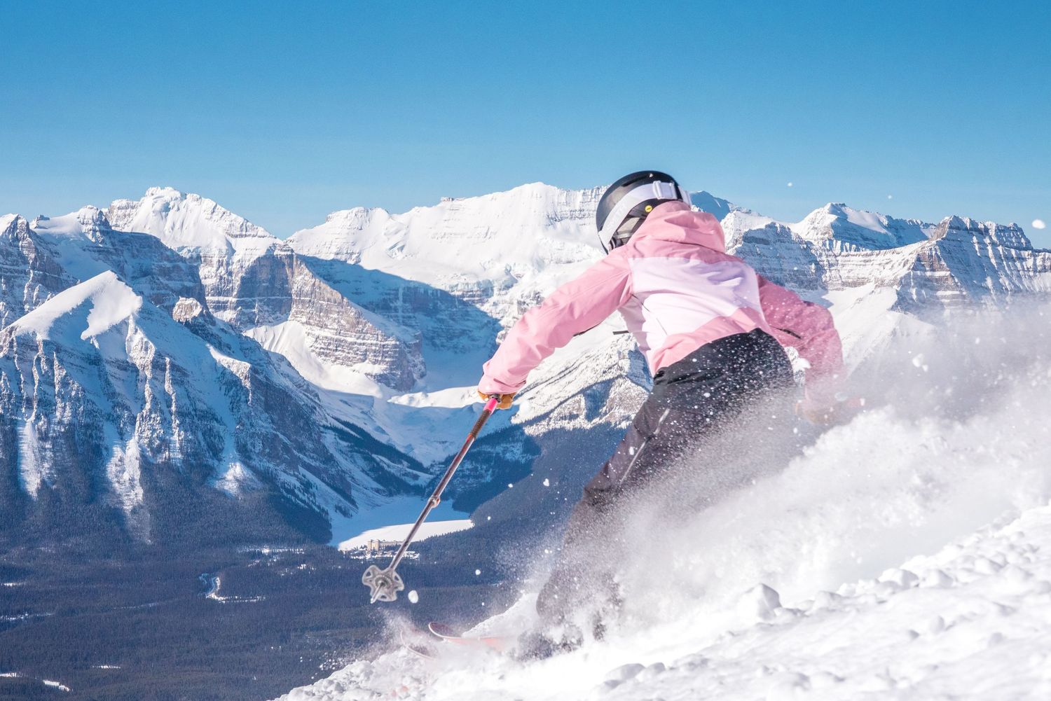 A skier in pink jacket performs a dynamic jump against a backdrop of snow-covered mountain peaks on a sunny day.