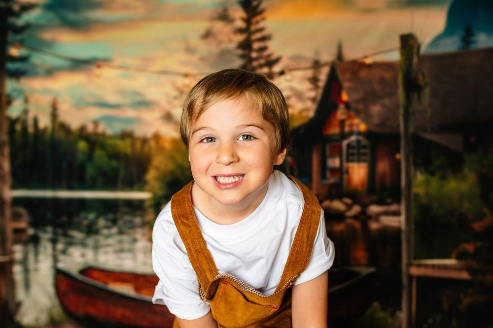 A smiling child in a white shirt and brown overalls poses in front of a lakeside cabin at sunset.