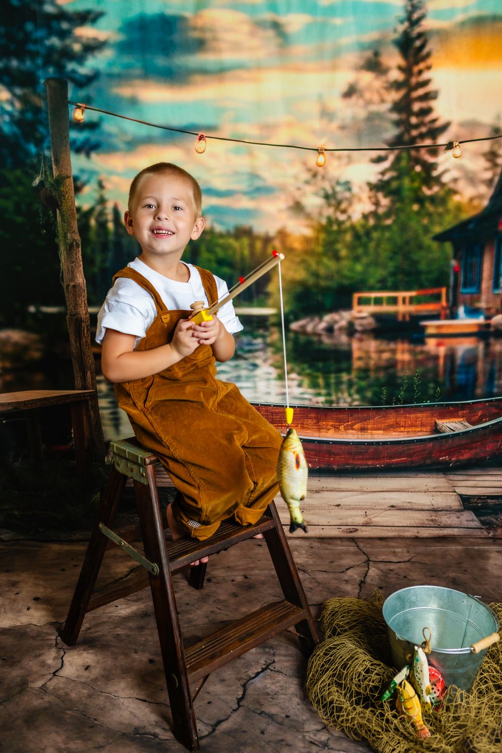 A child in brown overalls sits in a wooden chair by a lake during golden hour with fishing equipment nearby.